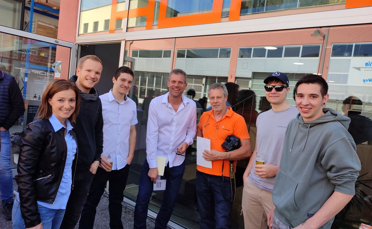 Gruppe von sechs Personen steht vor einem Gebäude mit orangefarbener Beschriftung. Einige halten Getränke in der Hand.